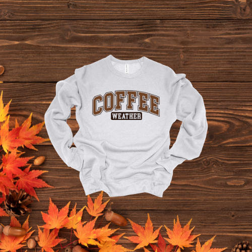 A heathered mustard colored crewneck sweatshirt with 'COFFEE WEATHER' text on the front, placed on a wooden surface with autumn leaves around.