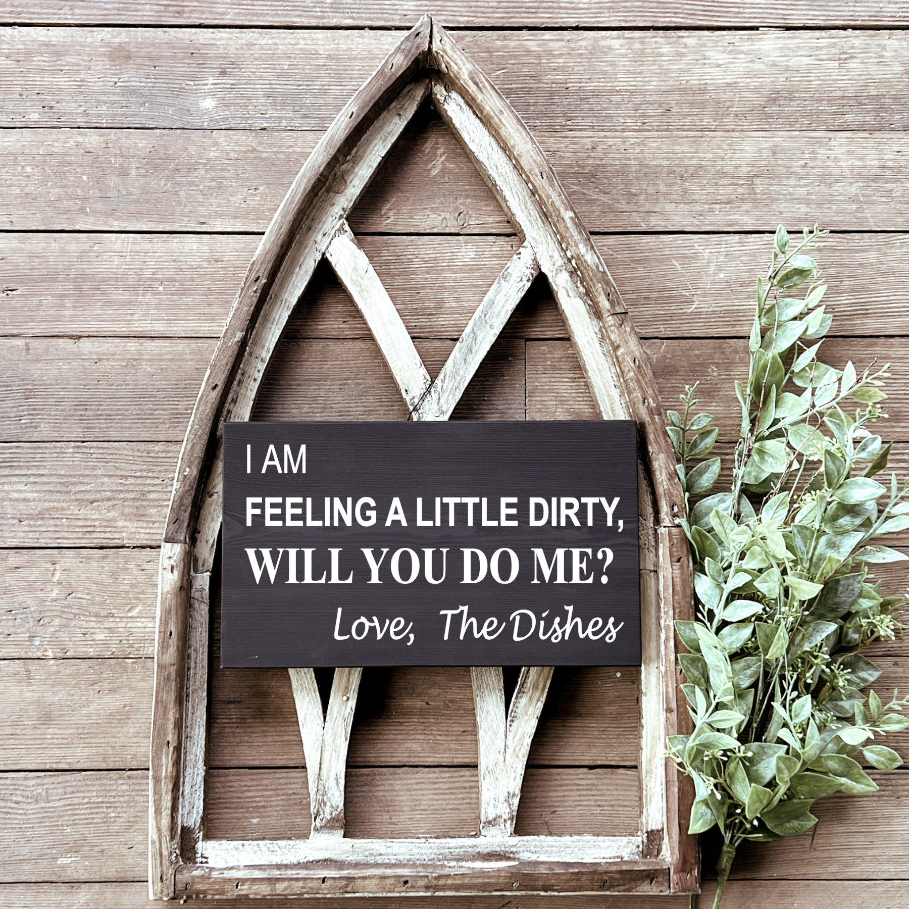 A humorous wooden sign with the phrase 'I am feeling a little dirty, will you do me? Love, The Dishes' written in black, placed in a rustic wooden frame, with some greenery to the side.