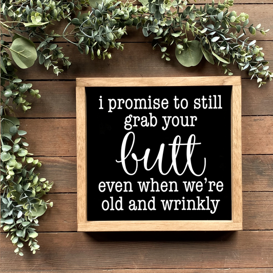 A framed wooden sign with the phrase 'I promise to still grab your butt even when we're old and wrinkly' written in white, displayed on a wooden surface with greenery around it.