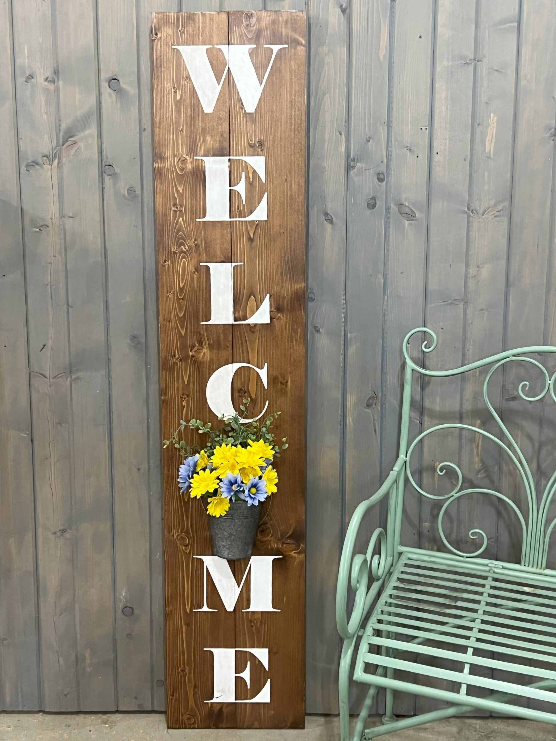 A wooden vertical 'WELCOME' sign with a flower planter built into the design, displayed against a grey wooden backdrop with a green metal chair to the side.