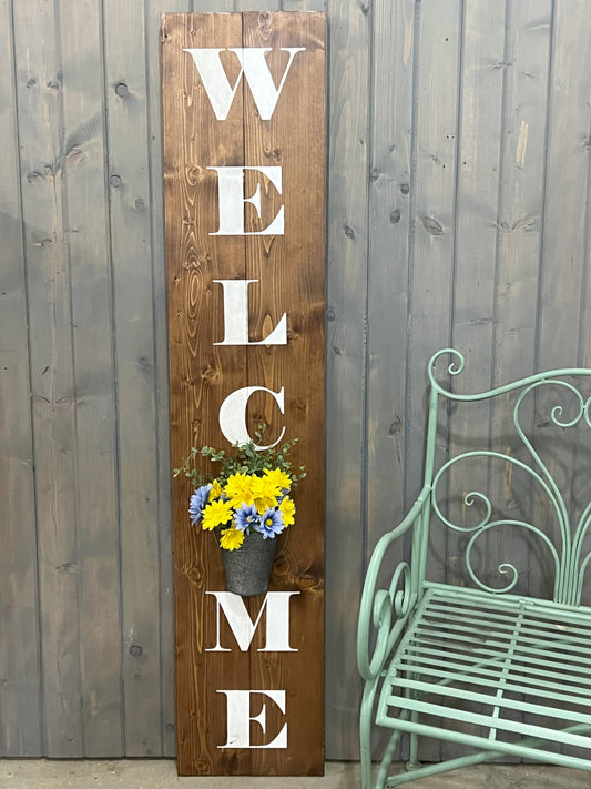 A wooden vertical 'WELCOME' sign with a flower planter built into the design, displayed against a grey wooden backdrop with a green metal chair to the side.