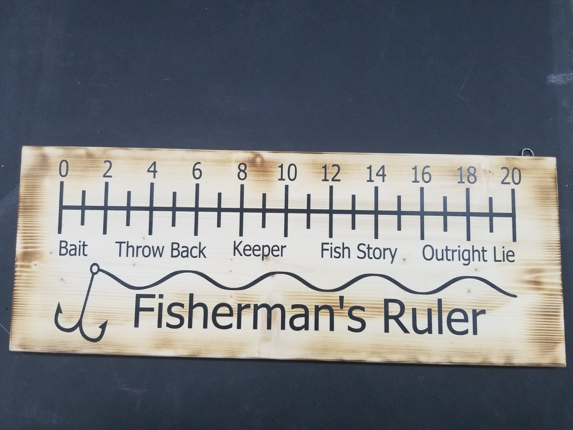 A wooden ruler sign with fishing-related measurements and terms such as 'THROAT', 'REEL', 'BAIT', 'CATCH', 'THROWBACK', 'BACK', 'KEEP', and 'FISH STORY' printed on it, along with the words 'FISHERMAN'S RULER' at the bottom.