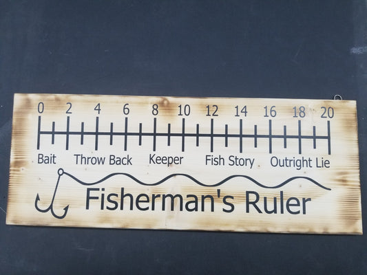 A wooden ruler sign with fishing-related measurements and terms such as 'THROAT', 'REEL', 'BAIT', 'CATCH', 'THROWBACK', 'BACK', 'KEEP', and 'FISH STORY' printed on it, along with the words 'FISHERMAN'S RULER' at the bottom.