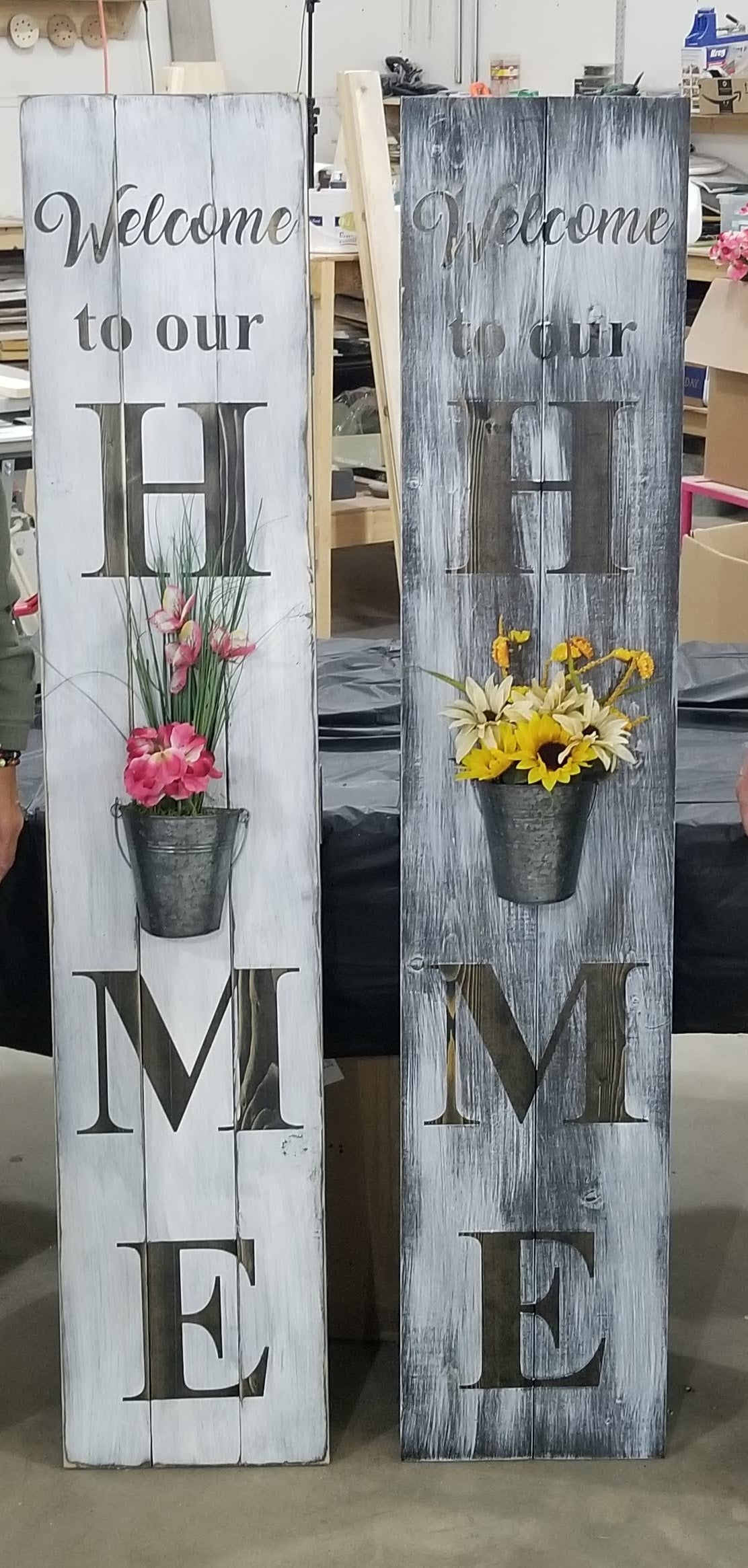 A wooden vertical 'WELCOME' sign with a flower planter built into the design, displayed against a grey wooden backdrop with a green metal chair to the side.