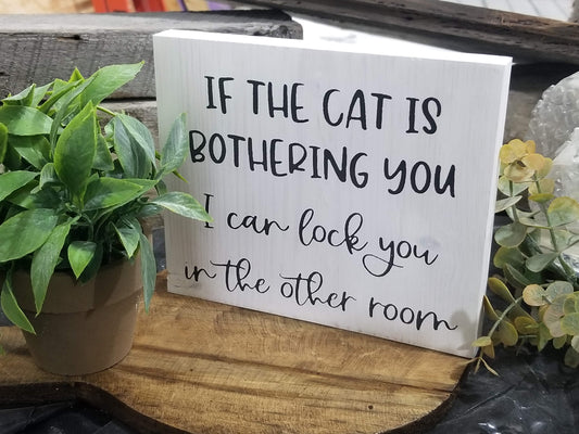 A white framed sign with black text that reads 'IF THE CAT IS BOthering YOU I CAN LOCK YOU IN THE OTHER ROOM', displayed on a wooden surface with a potted plant to the side.