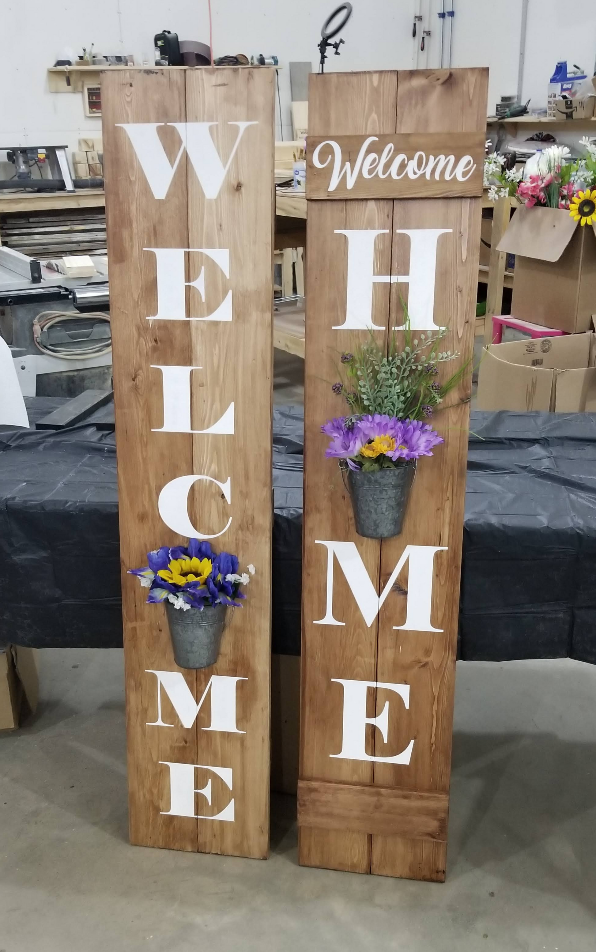 A wooden vertical 'WELCOME' sign with a flower planter built into the design, displayed against a grey wooden backdrop with a green metal chair to the side.