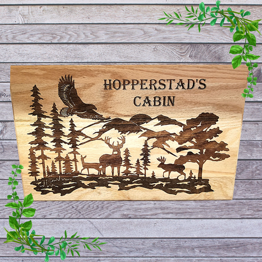 A custom engraved wooden sign featuring a wildlife scene with trees, mountains, and a cabin, surrounded by a forest.
