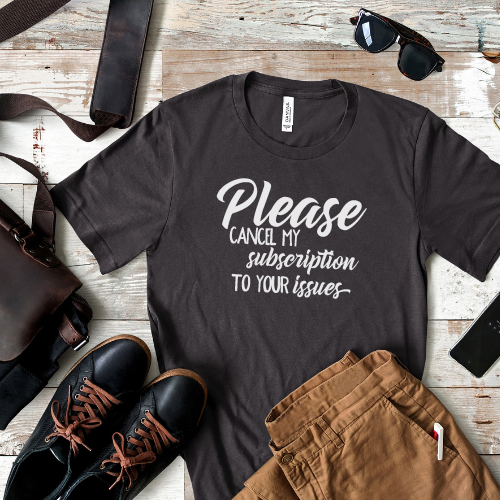 A gray t-shirt with the text 'Please cancel my subscription to your issues' printed on the front, displayed flat with a pair of brown pants, a pair of black shoes, a belt, sunglasses, and a leather bag.