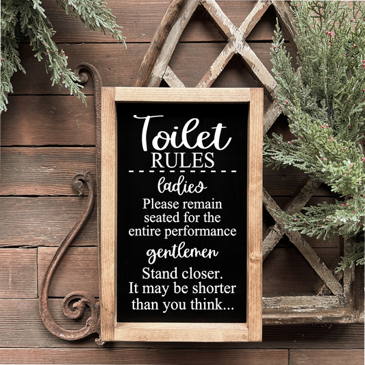 A wooden sign with a black or white background, humorous 'Toilet Rules' text, and a frame resembling a toilet bowl. It is positioned on a wall, likely in a home or office bathroom setting.