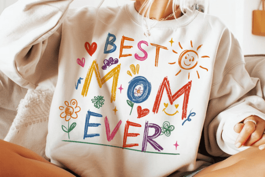 Cream-colored crewneck sweatshirt with "Best Mom Ever" written in colorful crayon-style letters surrounded by hearts, a sun, and flowers.