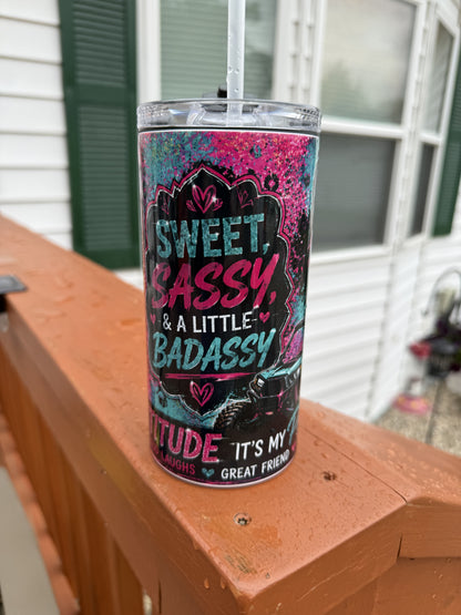 A clear plastic tumbler with a straw sits on a wooden deck railing in front of a house. The tumbler has a colorful graphic design with the text 'SWEET, BASSY, & A LITTLE BADASSY' and 'IT'S MY GREAT FRIEND'.
