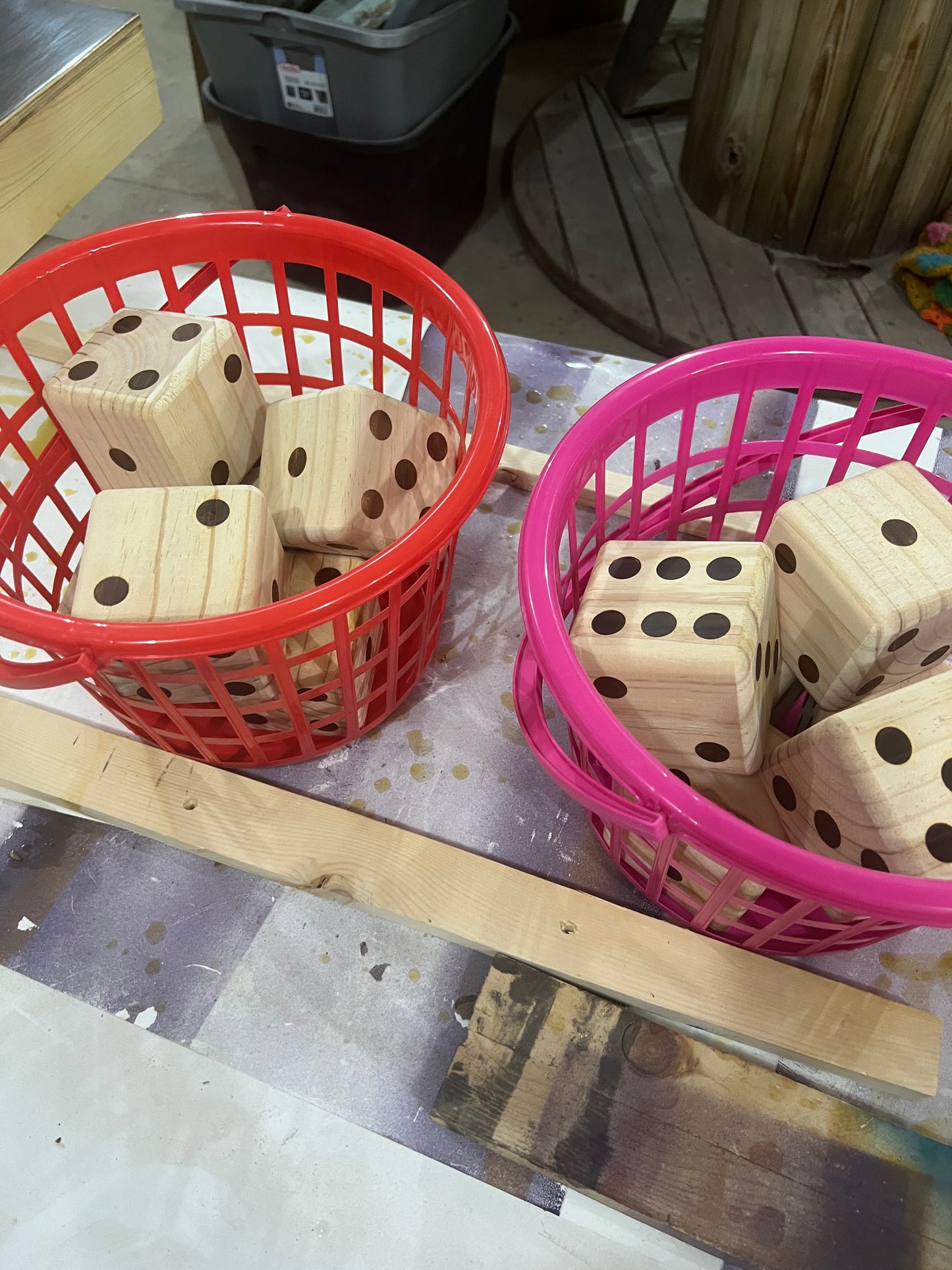 Two plastic baskets, one red and one pink, filled with unfinished wooden dice casting blocks on a workshop table.