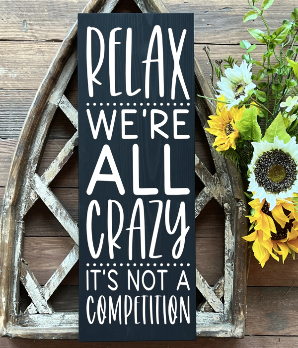 Wooden sign that says “Relax. We’re all crazy. It’s not a competition.” Available with white background and black text or black background and white text.