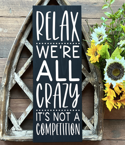 Wooden sign that says “Relax. We’re all crazy. It’s not a competition.” Available with white background and black text or black background and white text.