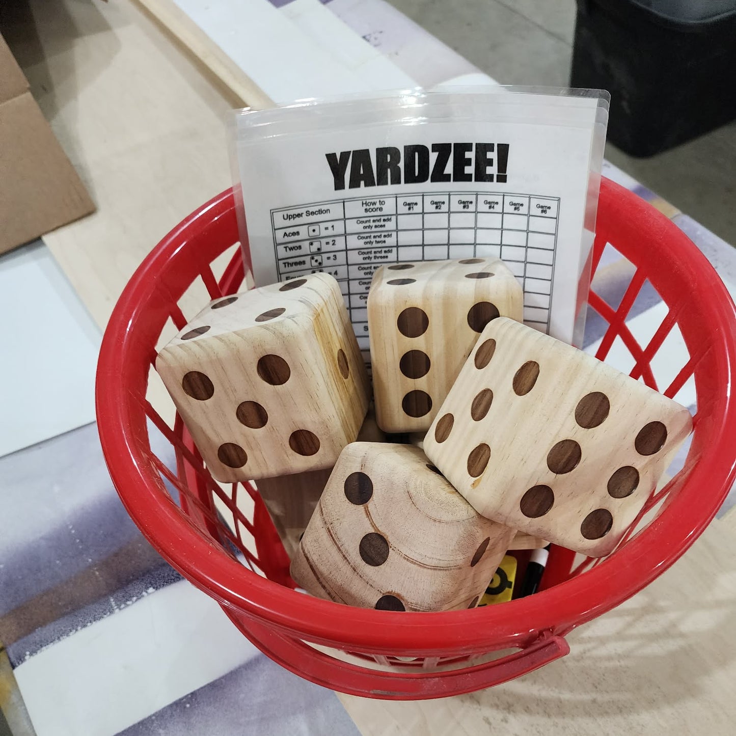 A red plastic basket containing four wooden yardzee dice and a yardzee score card, photographed on a tabletop in a real indoor setting.