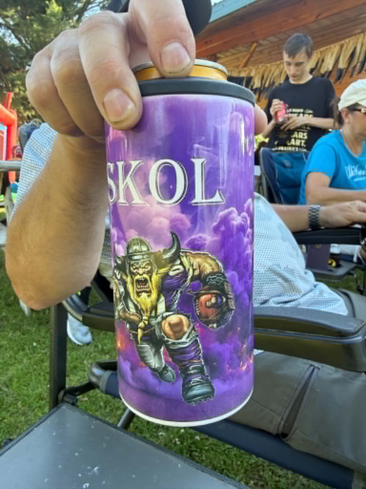 A hand holds a purple can of Skol beer outdoors at a gathering, featuring a graphic of a horned warrior and the word 'SKOL' in large letters.