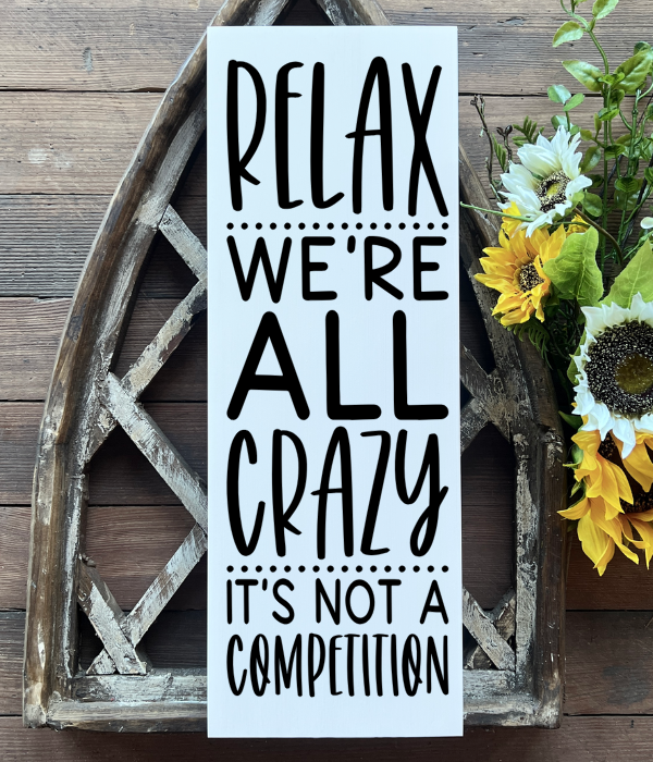 Wooden sign that says “Relax. We’re all crazy. It’s not a competition.” Available with white background and black text or black background and white text.