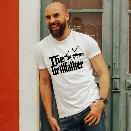 Man wearing a white t-shirt with 'The Grillfather' design, standing against a red and gray wall.